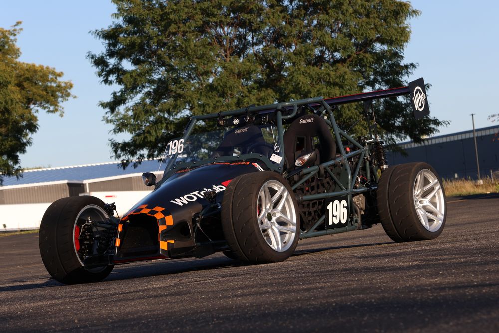 A low-angle view of the Goblin 196 kit car with exposed frame, sleek black body panels, and racing tires, parked on a paved lot under a blue sky, ready for action.