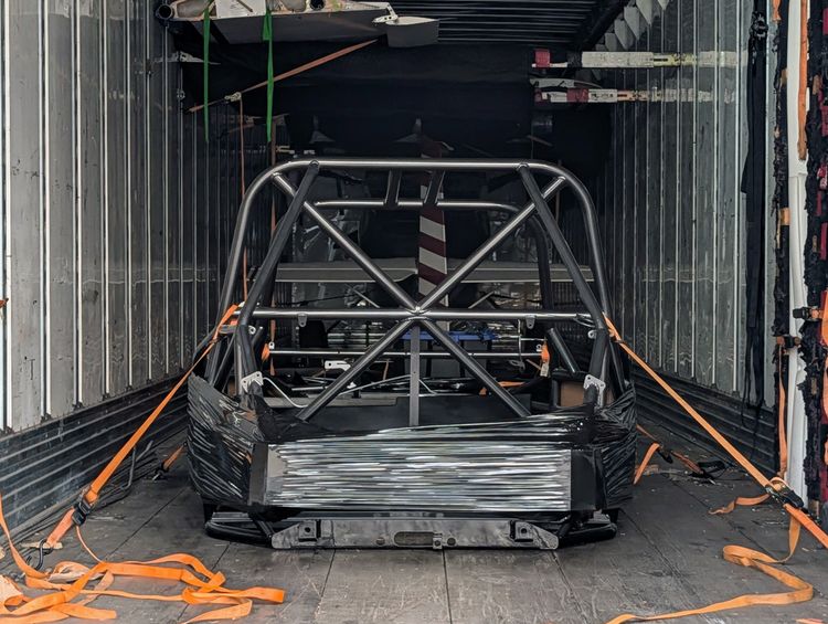 Goblin V2-006 chassis strapped down inside an enclosed trailer, viewed from the open rear doors, showing the T-Top main hoop with X-brace, standard door bars, and orange ratchet straps running to the trailer floor.