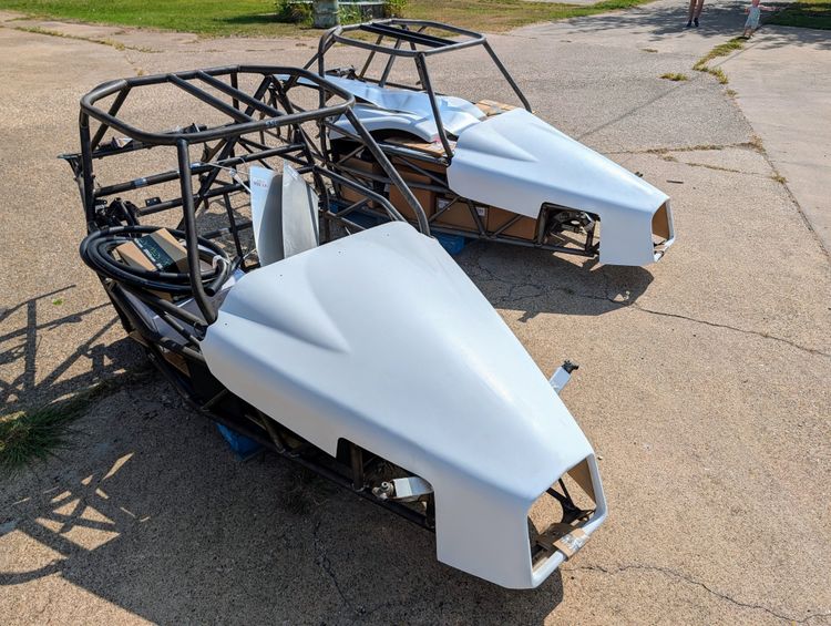 Two Goblin V1 full cage chassis staged outside the shop on pallets, both fitted with white unpainted hoods and loaded with boxed kit components and body panels.