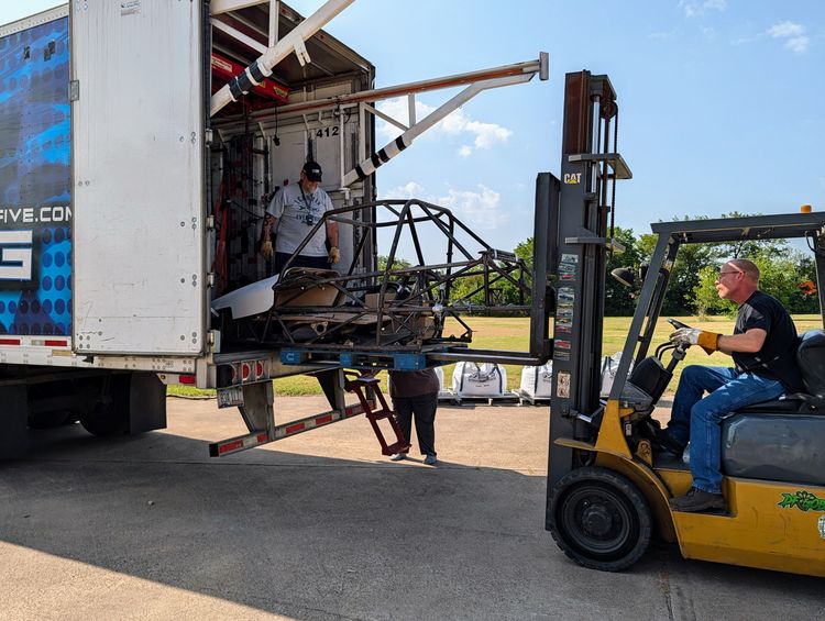 A forklift lifting a palletized Goblin V1 chassis into the back of a Cunningham Transport trailer, with the trailer's built-in crane boom extended overhead and a driver waiting inside to receive the load.