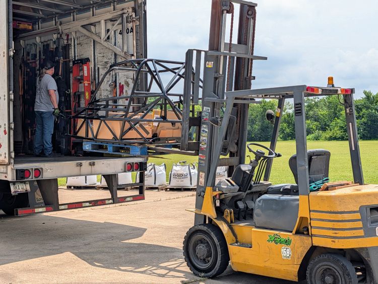 A Goblin V2 chassis in gloss black being loaded onto a Cunningham Transport trailer at the DF Kit Car shop, with shop staff inside the trailer guiding it into position.