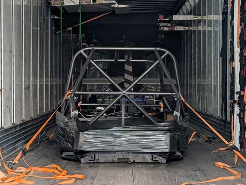 Goblin V2-006 chassis strapped down inside an enclosed trailer, viewed from the open rear doors, showing the T-Top main hoop with X-brace, standard door bars, and orange ratchet straps running to the trailer floor.